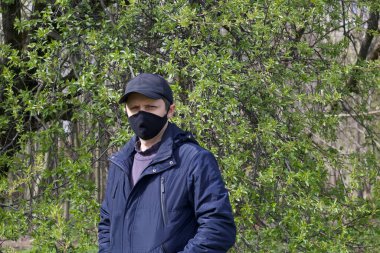 Bir bahar parkında yürüyen genç bir adamın portresi. Yüzünde siyah bir bez maske var. Bahar ceketi giymişti, şapkası kafasındaydı. Salgın sırasında temiz havada bir yürüyüş..