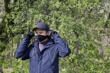Genç bir adam bir bahar parkında yürüyor ve jestler yapıyor. Yüzünde siyah bir bez maske var. Bahar ceketi giymişti, şapkası kafasındaydı. Salgın sırasında temiz havada bir yürüyüş..