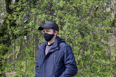 Bir bahar parkında yürüyen genç bir adamın portresi. Yüzünde siyah bir bez maske var. Bahar ceketi giymişti, şapkası kafasındaydı. Salgın sırasında temiz havada bir yürüyüş..