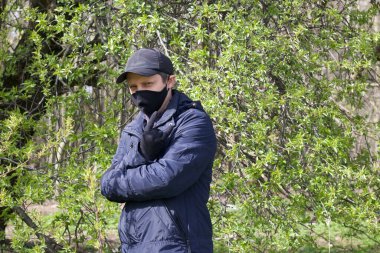 Genç bir adam bir bahar parkında yürüyor ve jestler yapıyor. Yüzünde siyah bir bez maske var. Bahar ceketi giymişti, şapkası kafasındaydı. Salgın sırasında temiz havada bir yürüyüş..