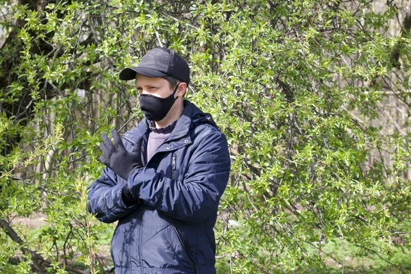 A young man walks through a spring park and gestures. On his face is a black cloth mask. Dressed in a spring jacket, cap on his head. A walk in the fresh air during a pandemic.