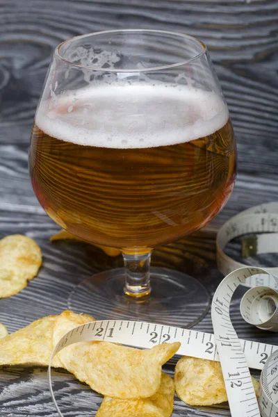 Glass of beer wrapped in measuring tape. Near potato chips. On brushed pine boards. Symbol of World No Diet Day.