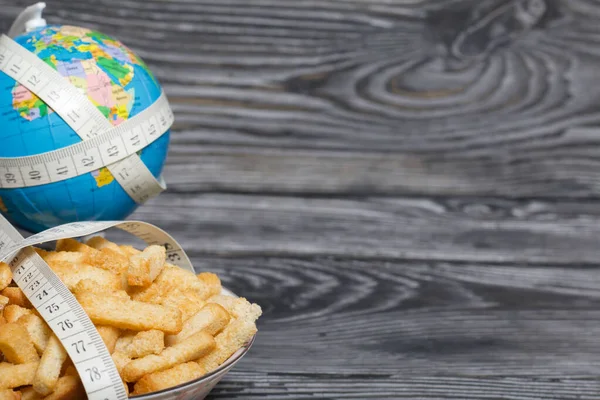 Globe wrapped with measuring tape. Near a cup with crackers. On brushed pine boards. Symbol of World No Diet Day.