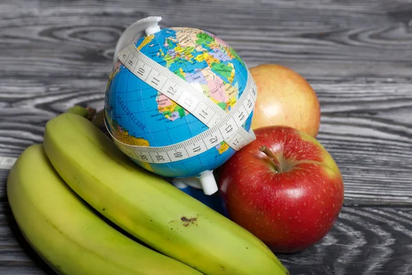 Globe wrapped with measuring tape. Near bananas and apples. On brushed pine boards. Symbol of World No Diet Day.