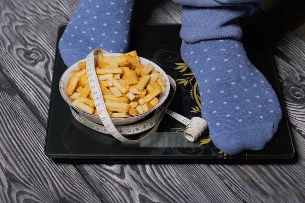 A man is standing on the floor scales. Near a cup with crackers for beer and a measuring tape. World No Diet Day. The scales stand on black brushed pine boards. Legs taken up close up.