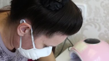 Koruyucu maskeli bir kız kendini soyutlayarak manikür yapar. Nazikçe tırnaklarına jöle sür. Yakınlarda manikür ve diğer aletler için bir lamba var. Yakın çekim..