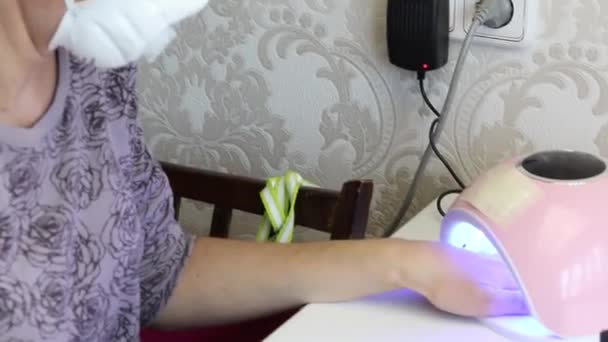 The girl in a protective mask dries gel polish in a lamp for drying. Self-isolation manicure. Close-up shot. — Stock Video