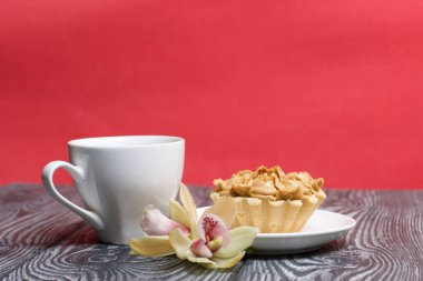 Kek ve sütlü bir fincan kahve. Yakınlarda orkide çiçekleri var. Pasta tereyağı kremasından yapılmış çiçeklerle süslenmiş. Kremanın karamel rengi var. Fırçalanmış çam ağaçlarında siyaha boyanmış.