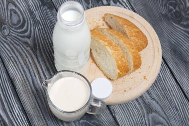 Bir şişe süt, bir bardak mayalanmış süt ve kesme tahtasındaki ciabatta ekmeği. Çam ağacından yapılmış tahtalarda.