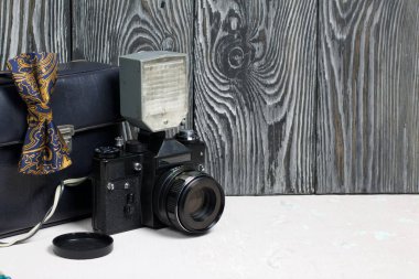 Fotoğrafçı aksesuarları. Kamera ve flaş. Kamerada bir papyon var. Fırçalanmış çam ağaçlarının arka planında.