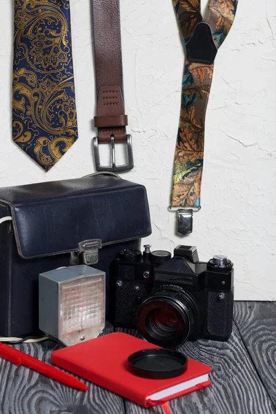 Photographer accessories. Camera and flash. Near tie, belt, suspenders, notebook and pen. On brushed pine boards.