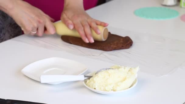 Auf einem Tisch wird die Lebensmittelfolie entwickelt. Eine Frau rollt mit einem Nudelholz einen braunen Teigklumpen. Zutaten und Werkzeuge für das Dessert. — Stockvideo