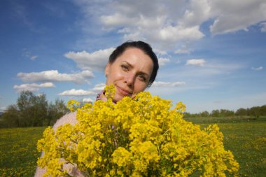 Sarı tecavüz buketi olan bir kız. Bahar çayırında. Mavi gökyüzünde kabarık bulutlar.