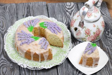 Lavantalı kek. Şeker kaplı. Lavanta rengi çiçeklerle süslenmiş. Küçük bir parça kesilir. Yakınlarda bir çaydanlık var..
