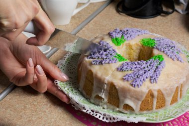 Lavantalı kek. Şeker kaplı. Lavanta rengi çiçeklerle süslenmiş. Bir kadın ondan küçük bir parça kesiyor..