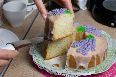 Lavantalı kek. Şeker kaplı. Lavanta rengi çiçeklerle süslenmiş. Bir kadın ondan küçük bir parça kesiyor..