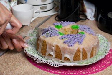 Lavantalı kek. Şeker kaplı. Lavanta rengi çiçeklerle süslenmiş. Bir kadın ondan küçük bir parça kesiyor..