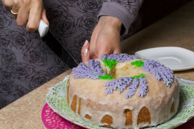 Lavantalı kek. Şeker kaplı. Lavanta rengi çiçeklerle süslenmiş. Bir kadın ondan küçük bir parça kesiyor..