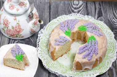 Lavantalı kek. Şeker kaplı. Lavanta rengi çiçeklerle süslenmiş. Küçük bir parça kesilir. Yakınlarda bir çaydanlık var..