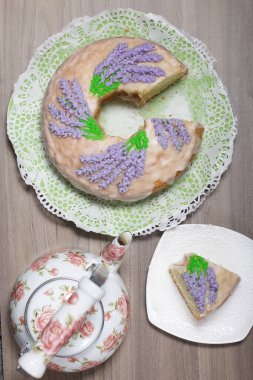 Lavantalı kek. Şeker kaplı. Lavanta rengi çiçeklerle süslenmiş. Küçük bir parça kesilir. Yakınlarda bir çaydanlık var..
