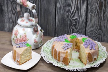 Lavantalı kek. Şeker kaplı. Lavanta rengi çiçeklerle süslenmiş. Küçük bir parça kesilir. Yakınlarda bir çaydanlık var..