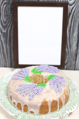 Lavantalı kek. Şeker kaplı. Lavanta rengi çiçeklerle süslenmiş. Yakınlarda boş bir fotoğraf çerçevesi var..