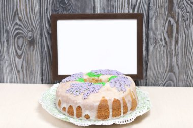 Lavantalı kek. Şeker kaplı. Lavanta rengi çiçeklerle süslenmiş. Yakınlarda boş bir fotoğraf çerçevesi var..