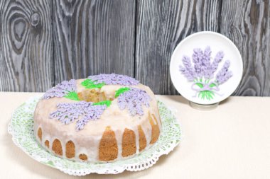 Lavantalı kek. Şeker kaplı. Lavanta rengi çiçeklerle süslenmiş. Cilalı bir tabağın yanında..