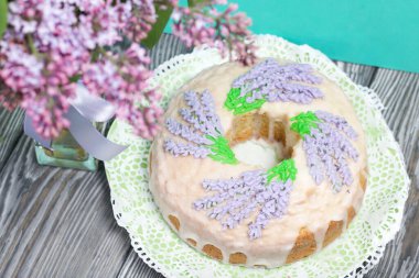 Lavantalı kek. Şeker kaplı. Lavanta rengi çiçeklerle süslenmiş. Bir vazonun yanında bir demet leylak..