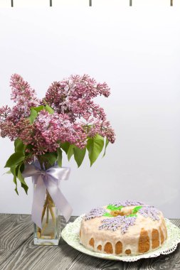 Lavantalı kek. Şeker kaplı. Lavanta rengi çiçeklerle süslenmiş. Bir vazonun yanında bir demet leylak..