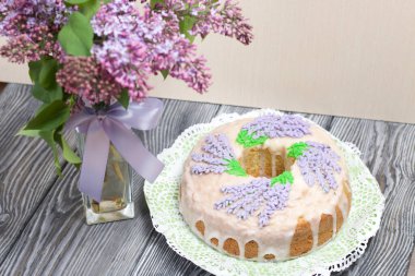 Lavantalı kek. Şeker kaplı. Lavanta rengi çiçeklerle süslenmiş. Bir vazonun yanında bir demet leylak..