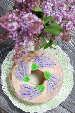 Lavantalı kek. Şeker kaplı. Lavanta rengi çiçeklerle süslenmiş. Bir vazonun yanında bir demet leylak..