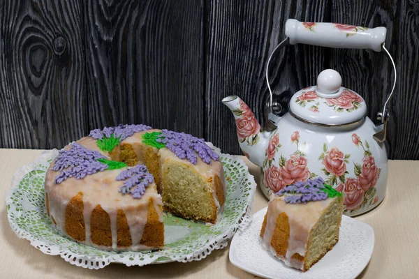 Lavantalı kek. Şeker kaplı. Lavanta rengi çiçeklerle süslenmiş. Küçük bir parça kesilir. Yakınlarda bir çaydanlık var..