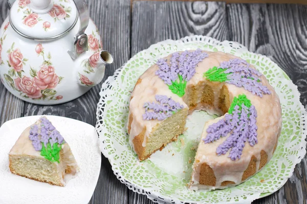 Lavantalı kek. Şeker kaplı. Lavanta rengi çiçeklerle süslenmiş. Küçük bir parça kesilir. Yakınlarda bir çaydanlık var..