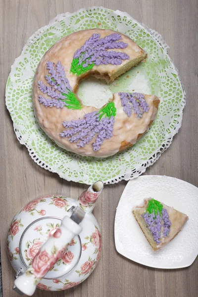 Lavantalı kek. Şeker kaplı. Lavanta rengi çiçeklerle süslenmiş. Küçük bir parça kesilir. Yakınlarda bir çaydanlık var..