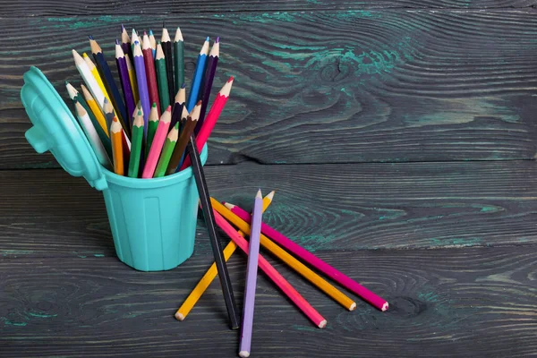 Pencil filled with colored pencils. In the form of a garbage container. Near a few pencils. Against the background of brushed pine boards painted in black and green.