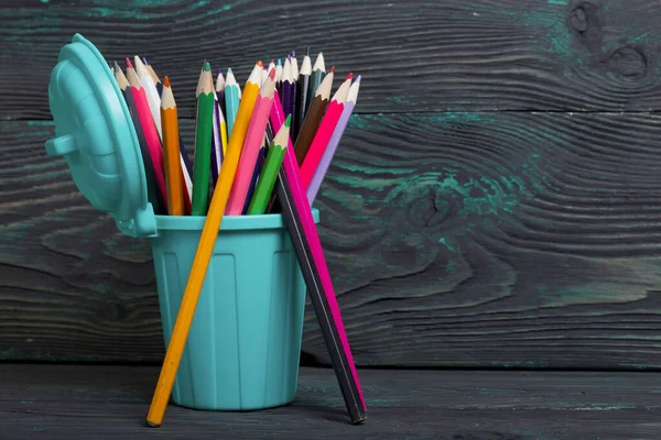 Pencil filled with colored pencils. In the form of a garbage container. Near a few pencils. Against the background of brushed pine boards painted in black and green.