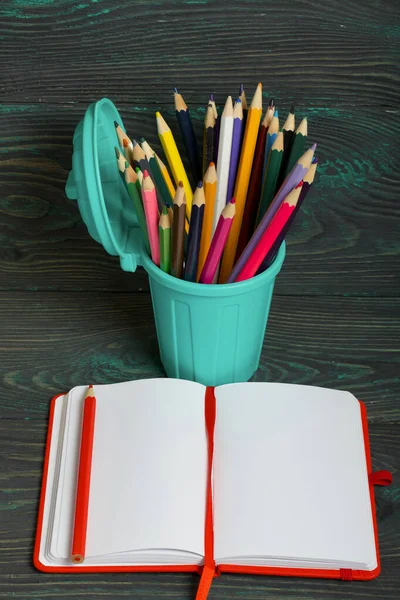 Pencil filled with colored pencils. In the form of a garbage container. Near an open notebook. Against the background of brushed pine boards painted in black and green.