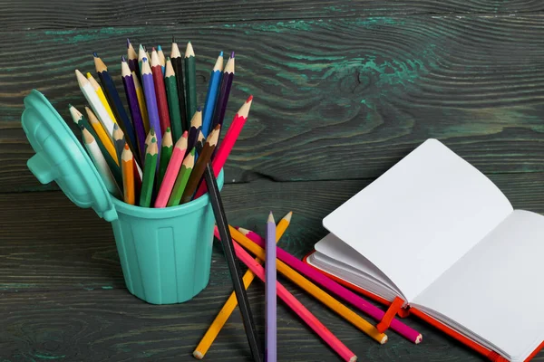 Pencil filled with colored pencils. In the form of a garbage container. Nearby is an open notebook and several pencils. Against the background of brushed pine boards painted in black and green.