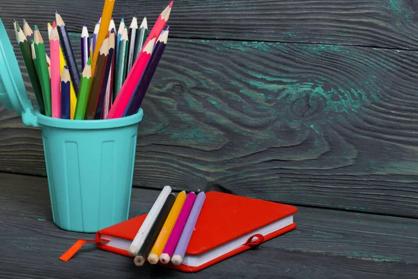 Pencil filled with colored pencils. In the form of a garbage container. Near a notebook and some pencils. Against the background of brushed pine boards painted in black and green.