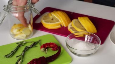 Bir kadın cam kavanoza limon dilimleri koyup tuz serpiyor. Biberiyenin yanında, tuz ve acı biber. Limon tuzlamak için gerekli malzemeler.