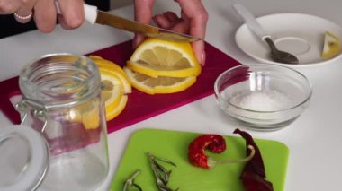 Kadın dilimleme tahtasındaki limonu dilimliyor. Biberiyenin yanında, tuz ve acı biber. Limon tuzlamak için gerekli malzemeler