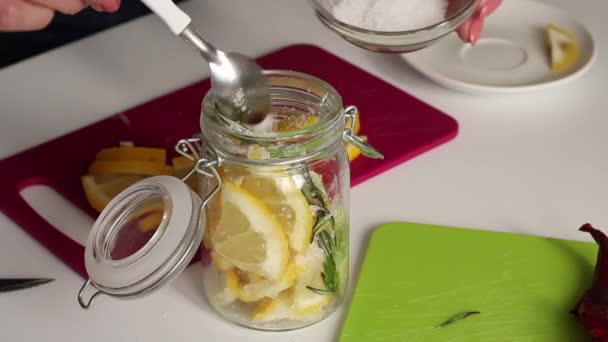 Een vrouw voegt zout toe aan citroenschijfjes, rozemarijn en hete peper in een glazen pot. Mengt ingrediënten van citroenzout. — Stockvideo