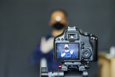 Koruyucu maskeli bir blogcu kamerada kendi fotoğrafını çeker. Kamerasına ve üzerindeki görüntüye odaklanmış durumdalar. Diğer her şey bulanık..