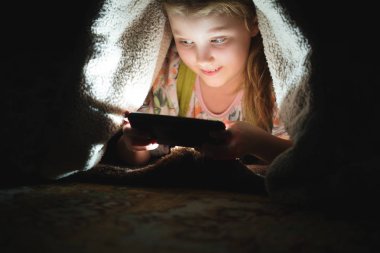 Kız gece battaniyenin altında yerde akıllı bir telefonla oyun oynuyor.
