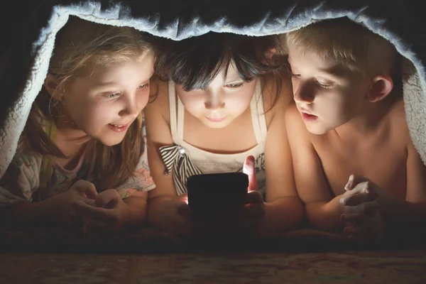 Kinderen spelen 's nachts spelletjes op een smartphone onder deken op de vloer — Stockfoto