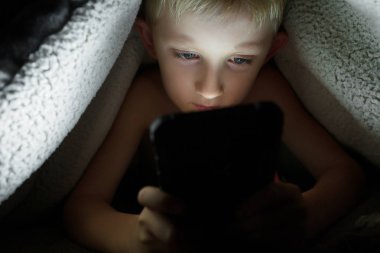 Çocuk gece akıllı bir telefonla yerde battaniyenin altında oyun oynuyor.