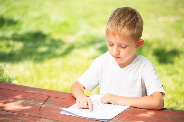 白いTシャツを着た小さな男の子が学校の概念に戻って屋外で読書しています. — ストック写真