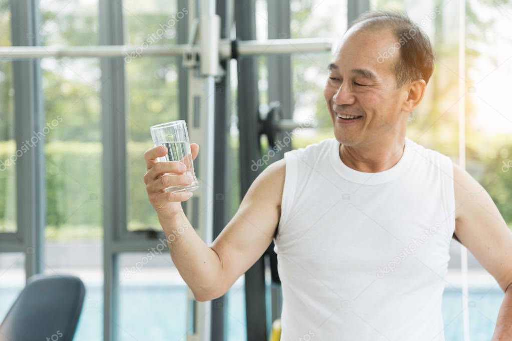 Asian senior man holding glass of water at a gym. Tired, thirsti