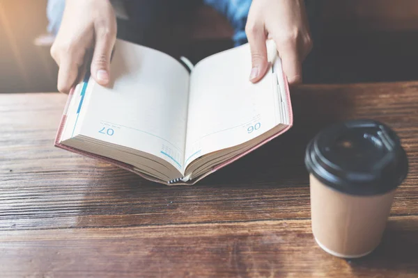 Focus on book. Woman holding notebook with a cup of coffee on wo — Stock Photo, Image